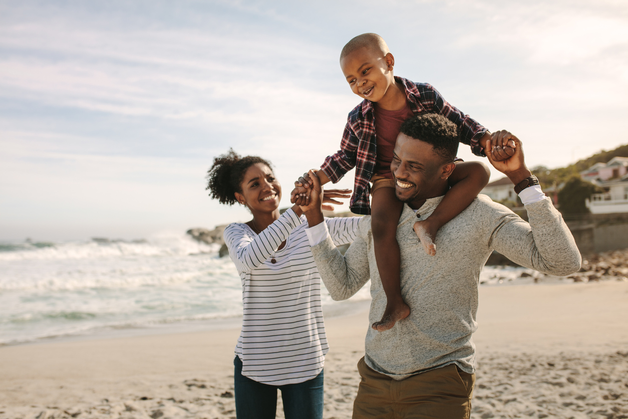 Parents carrying son on shoulders on beach vacation, following summer health safety tips