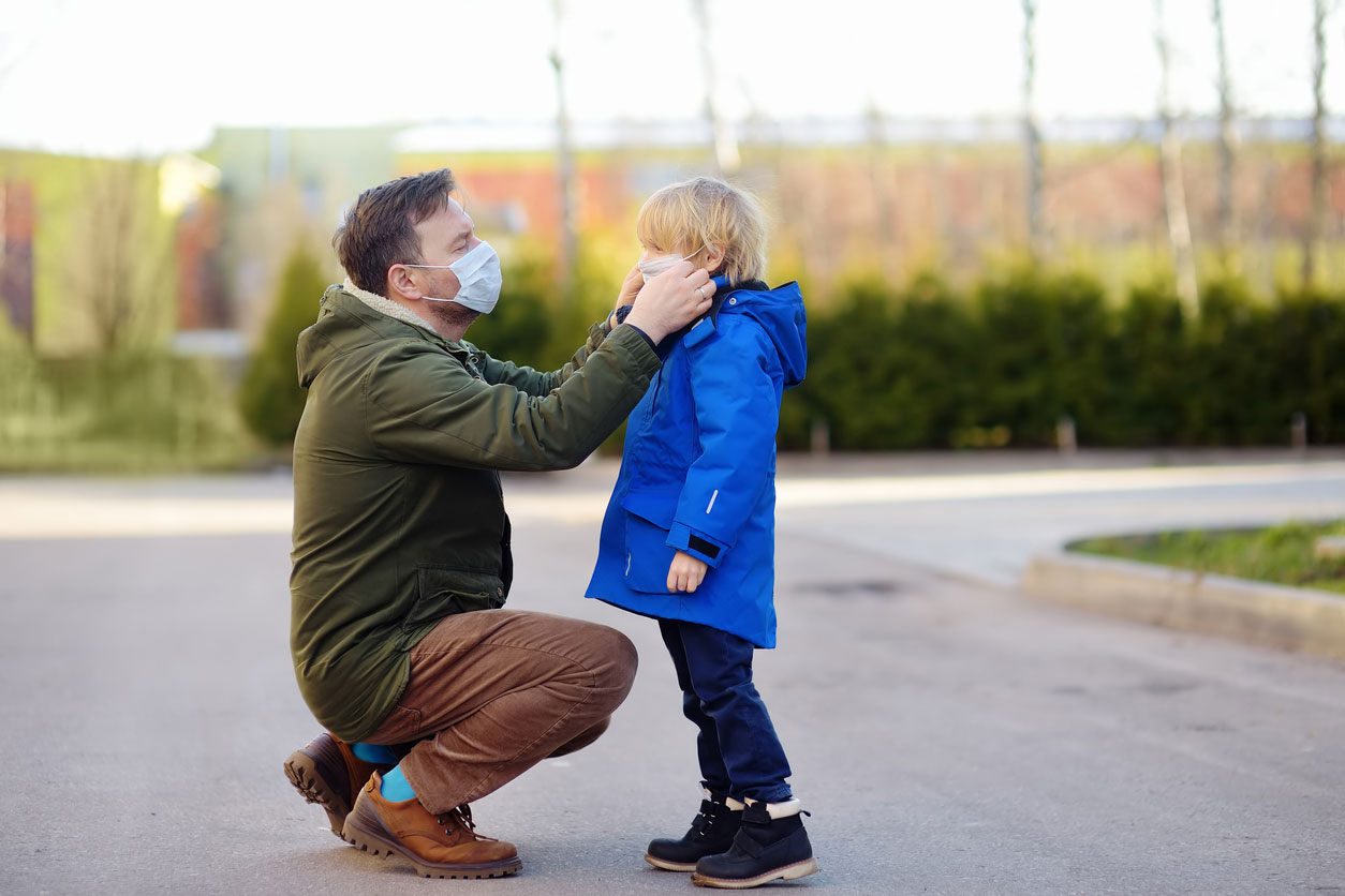 Mature man wearing a protective mask puts a face mask on a his son in airport, supermarket or other public place. Safety during COVID-19 outbreak. Epidemic of virus covid. Discussing the common covid-19 myths.